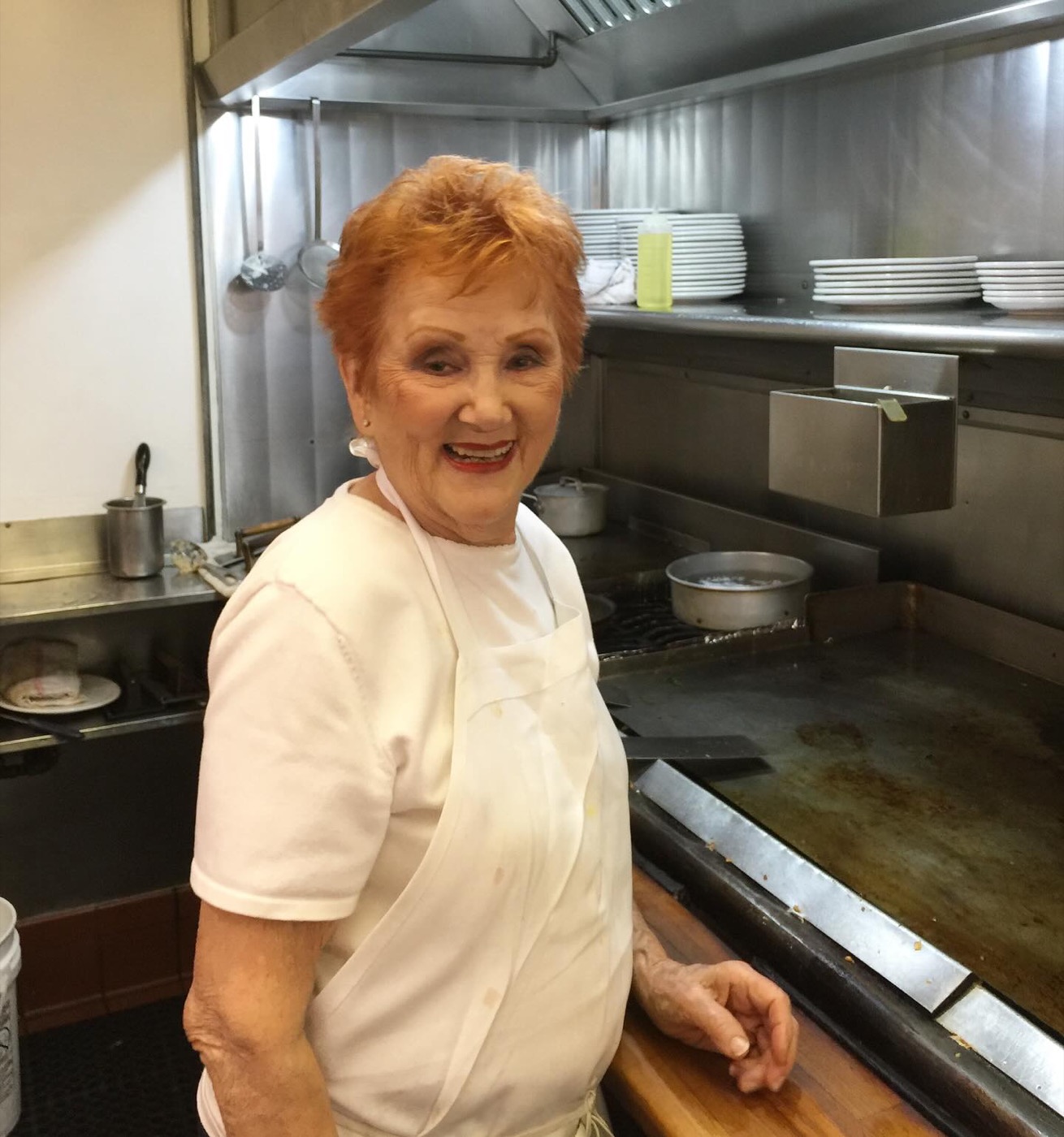 Charlie Sue Gilbert, smiling in a white apron, standing at the grill.